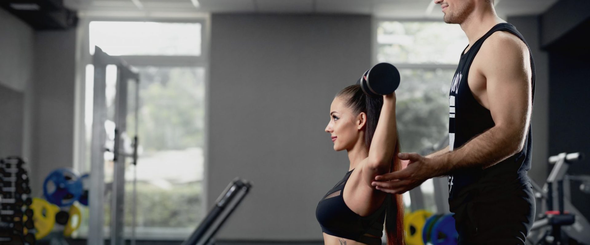 male-personal-trainer-helping-woman-working-with-heavy-dumbbells-gym-scaled
